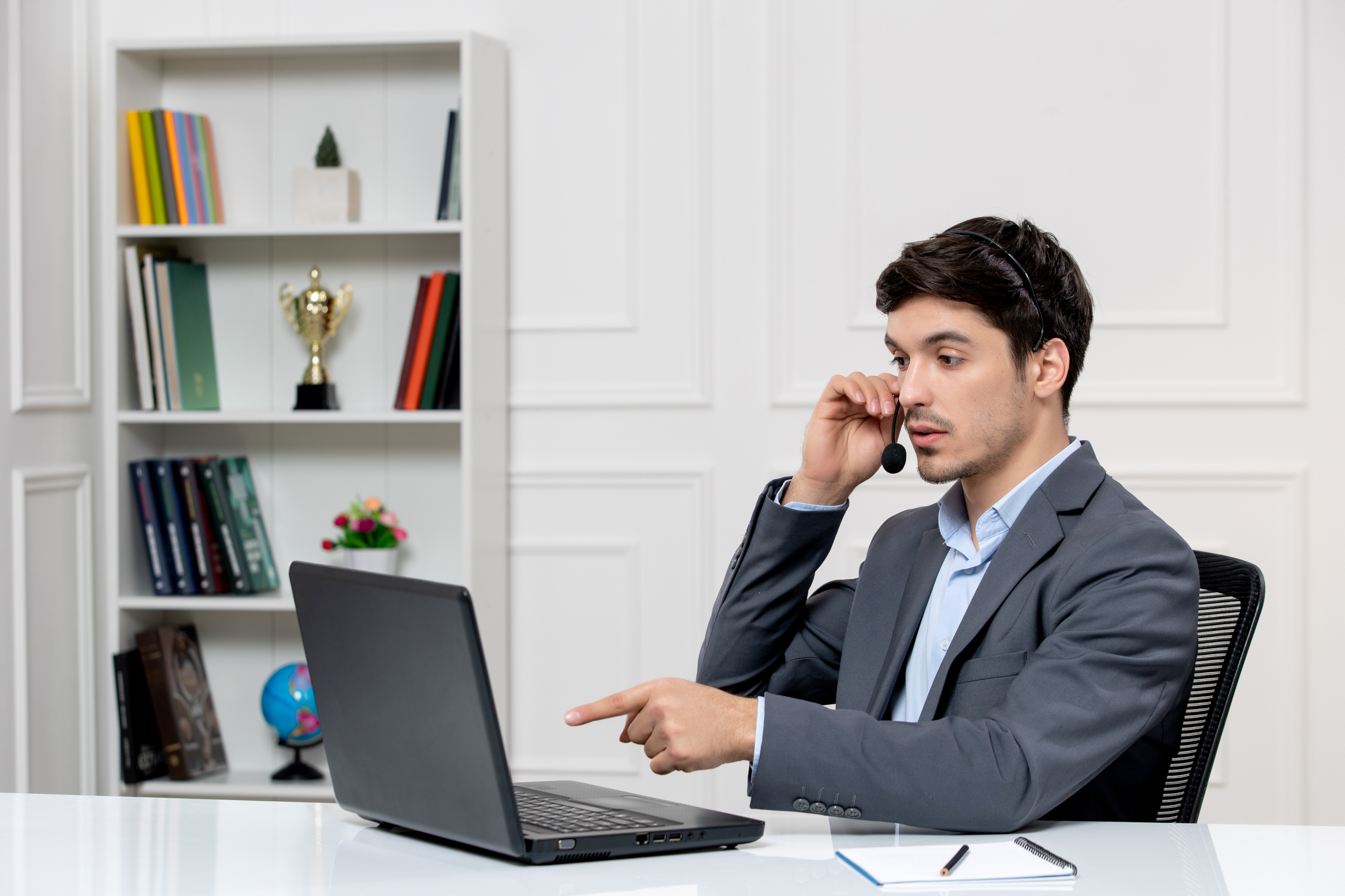 Male in grey suit with computer and headset pointing at screen